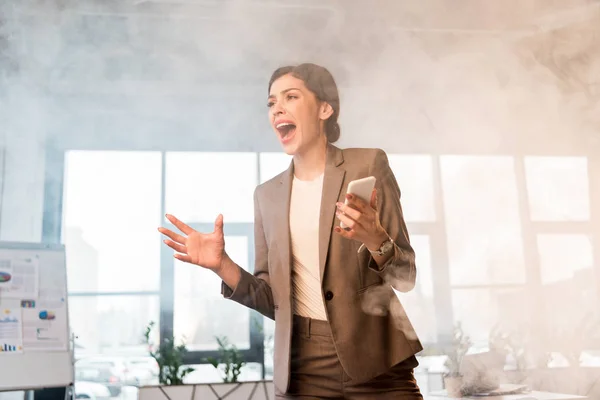 Terrifié femme d'affaires hurlant Whle tenant smartphone dans le bureau avec de la fumée — Photo de stock