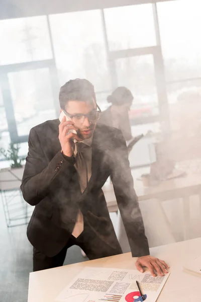 Scared businessman in glasses talking on smartphone in office with smoke near coworker — Stock Photo