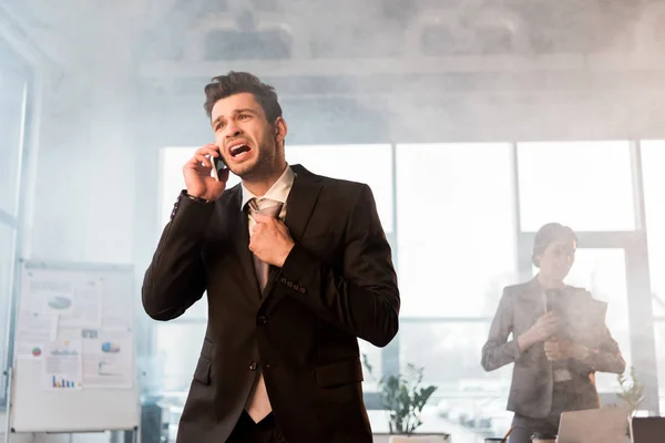 Terrified businessman talking on smartphone in office with smoke near coworker — Stock Photo