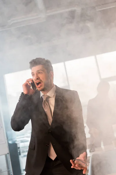 Испуганный бизнесмен разговаривает по смартфону и кричит в офисе с дымом рядом с коллегой — стоковое фото