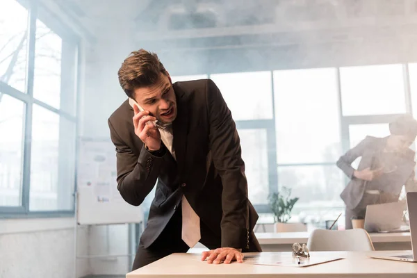 Испуганный бизнесмен разговаривает на смартфоне в офисе с дымом рядом с коллегой-женщиной — стоковое фото