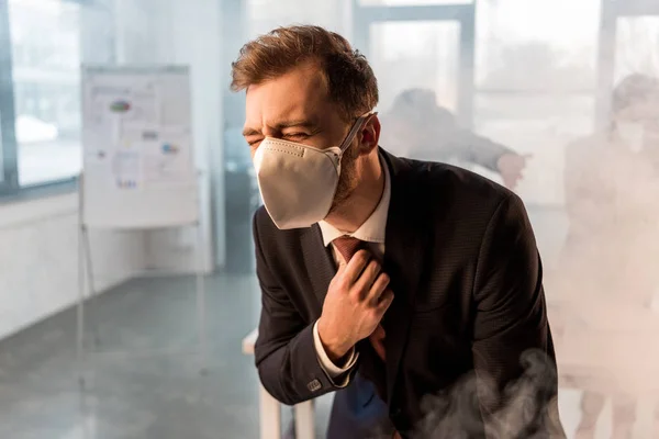 Enfoque selectivo de hombre de negocios en la máscara de pie en la oficina con humo cerca de compañeros de trabajo - foto de stock