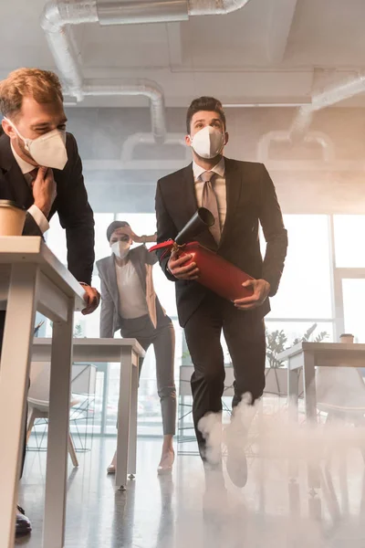 Businessman in mask holding extinguisher near coworkers in office with smoke — Stock Photo