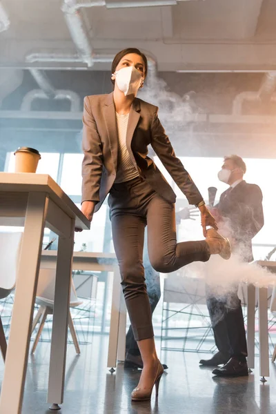 Mujer de negocios en la máscara que sostiene el zapato mientras está de pie cerca de colegas en la oficina con humo - foto de stock