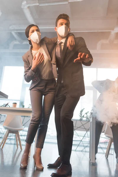 Hombre de negocios en la máscara de pie cerca de una compañera de trabajo en la oficina con humo - foto de stock