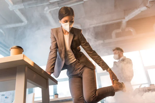 Mujer en máscara sosteniendo zapato mientras está de pie cerca de compañeros de trabajo en la oficina con humo - foto de stock