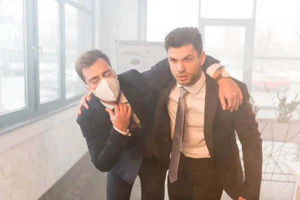 Terrified businessman in mask standing with serious coworker in office with smoke — Stock Photo