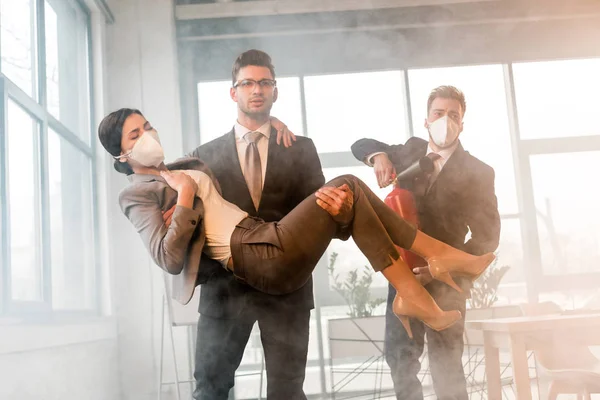 Guapo hombre de negocios en gafas sosteniendo en brazos a compañera de trabajo en máscara cerca de colega sosteniendo extintor en oficina con humo - foto de stock