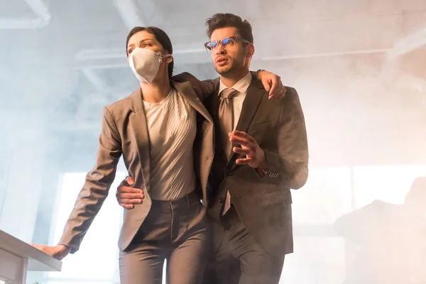 Bel homme d'affaires dans des lunettes debout avec une collègue femme portant un masque au bureau avec de la fumée — Photo de stock