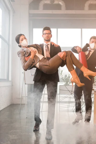 Bonito empresário em óculos segurando em braços fêmea colega de trabalho com olhos fechados perto colega segurando extintor no escritório com fumaça — Fotografia de Stock