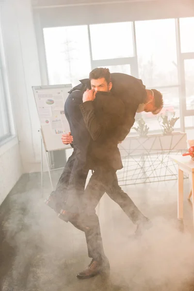 Terrified businessman holding in arms coworker in mask while covering nose in office with smoke — Stock Photo