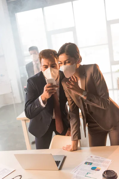 Selective focus of terrified colleagues in masks looking at smartphone in office with smoke — Stock Photo