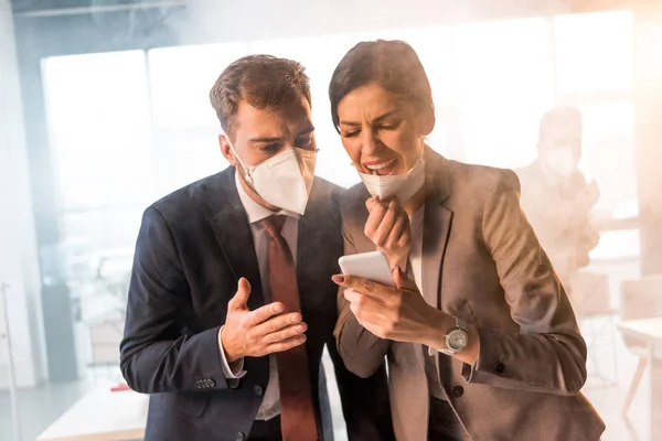Foco seletivo de mulher aterrorizada gritando perto colega na máscara enquanto olha para o smartphone no escritório com fumaça — Fotografia de Stock