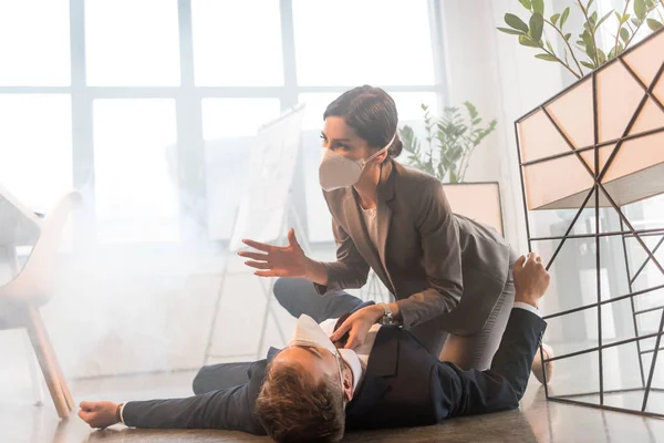 Verängstigte Geschäftsfrau mit Maske sitzt neben Kollegin, die im Büro mit Rauch am Boden liegt — Stockfoto