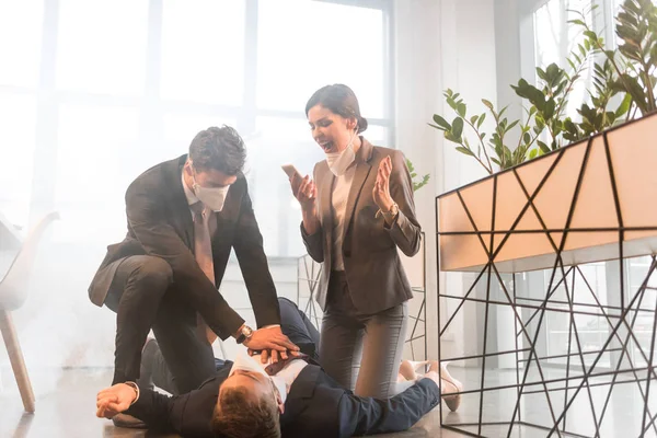 Terrified businesswoman screaming near colleague lying on floor while man giving first aid  in office with smoke — Stock Photo