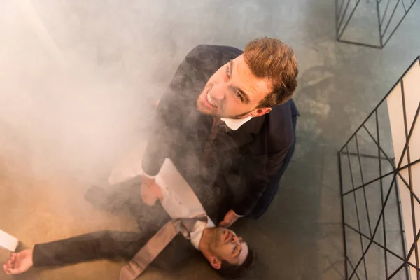 Vue aérienne d'un homme d'affaires effrayé assis sur le sol près d'un collègue les yeux fermés au bureau avec de la fumée — Photo de stock