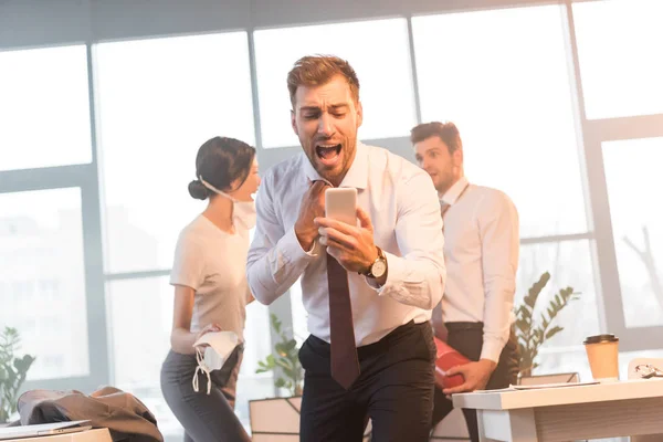 Enfoque selectivo del hombre de negocios asustado gritando mientras mira el teléfono inteligente con compañeros de trabajo en la oficina con humo en el fondo - foto de stock