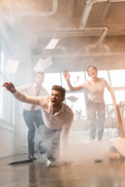 Terrified businessman falling on floor while running and yelling near coworkers in office with smoke — Stock Photo