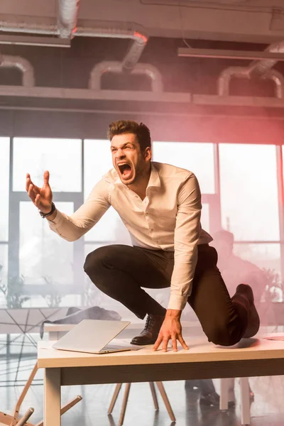 Scared businessman screaming while sitting on desk with laptop near coworker in office with smoke — Stock Photo