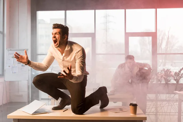 Terrified businessman screaming while sitting on desk with laptop near coworkers in office with smoke — Stock Photo