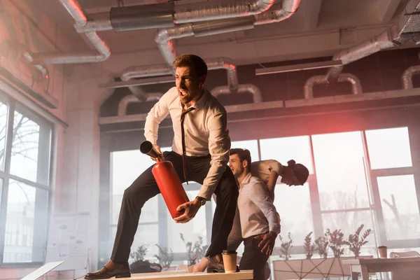 Verängstigter Geschäftsmann brüllt, während er Feuerlöscher in der Hand hält und neben Kollegen im Büro mit Rauch auf dem Schreibtisch steht — Stockfoto