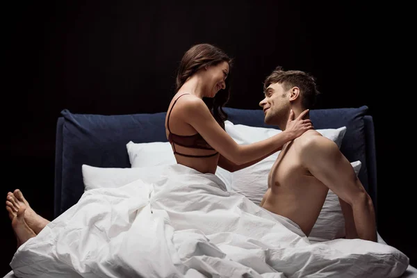 Улыбающаяся женщина в сексуальном нижнем белье обнимающая мужчину в постели изолированная на черном — стоковое фото