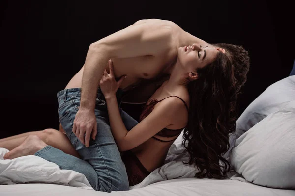 Hombre sin camisa besando hermosa mujer en cama aislado en negro - foto de stock
