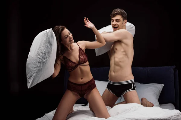 Счастливая молодая сексуальная пара борется с подушками на кровати изолированы на черном — стоковое фото