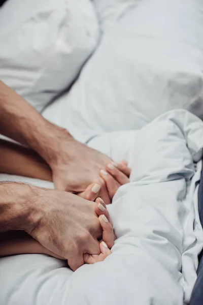 Vista recortada de la pareja cogida de la mano y acostada en la cama en casa con espacio para copiar - foto de stock