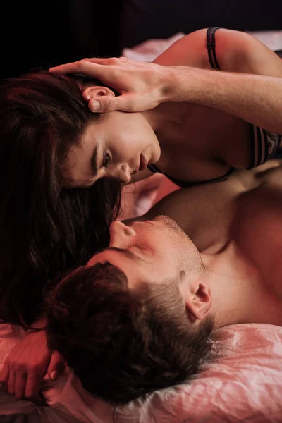 Mann umarmt sanft schöne junge Frau im Bett isoliert auf schwarz mit neonrotem Licht — Stockfoto