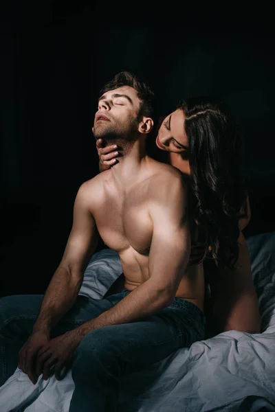 Beautiful young sexy woman touching neck of handsome man in bed isolated on black — Stock Photo