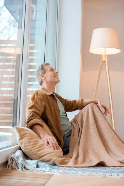 Uomo sorridente guardando in alto mentre seduto sul pavimento da grande finestra a nuova casa — Stock Photo