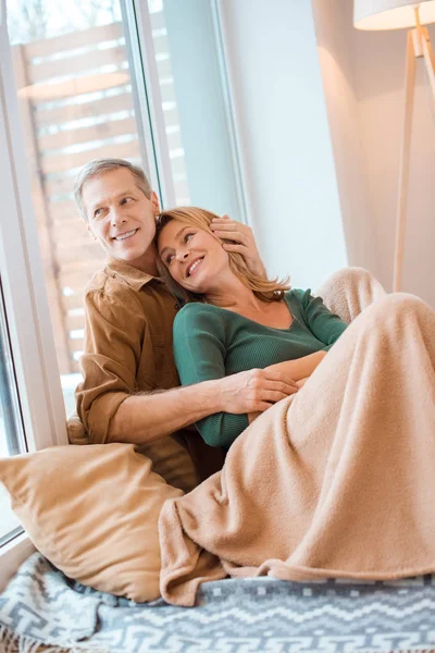 Couple souriant étreignant assis sur le sol par une grande fenêtre à la nouvelle maison — Photo de stock