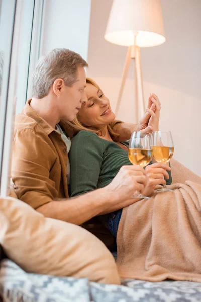 Happy couple holding glasses of white wine while resting under soft fleece blanket — Stock Photo