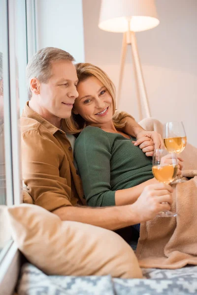 Coppia sorridente che abbraccia e tiene i calici di vino bianco mentre riposa sul pavimento di una nuova casa — Foto stock