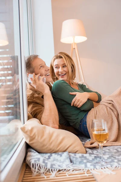 Sonriente hombre hablando en el teléfono inteligente mientras está sentado en el suelo con su esposa - foto de stock