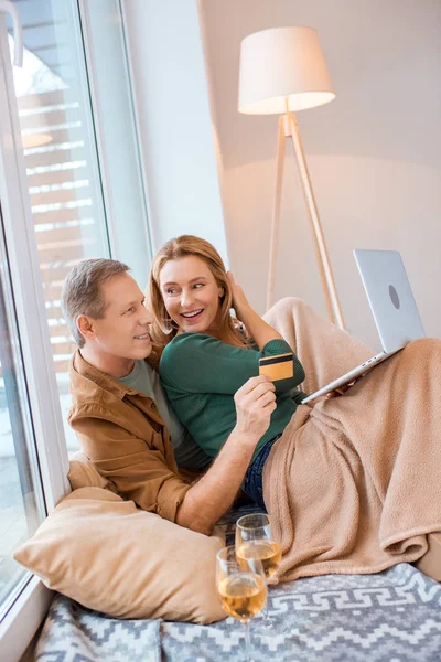 Sonriente hombre sosteniendo la tarjeta de crédito mientras usa el ordenador portátil con esposa - foto de stock