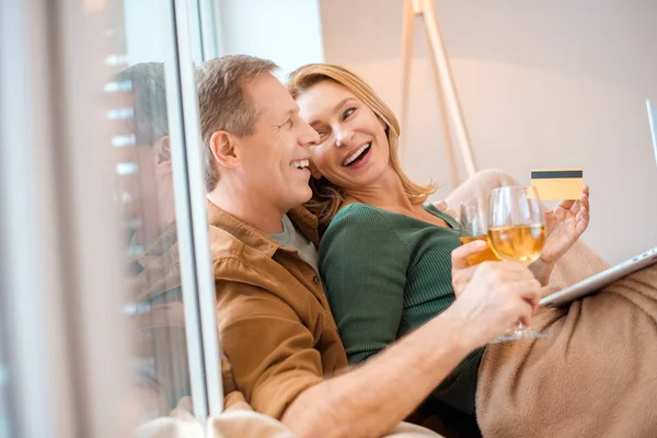 Ridere coppia che tiene i bicchieri di vino bianco mentre seduto alla finestra a casa — Foto stock