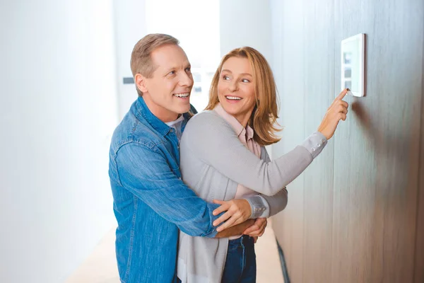 Feliz hombre abrazando esposa usando el panel de control del hogar inteligente - foto de stock