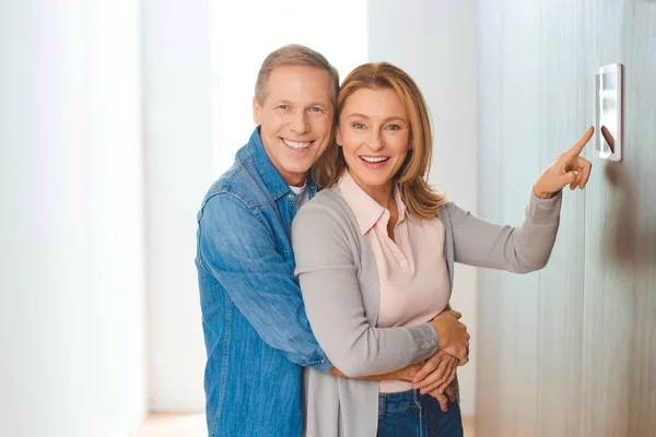 Усміхнений чоловік обіймає дружину, вказуючи на панель управління розумним будинком — стокове фото