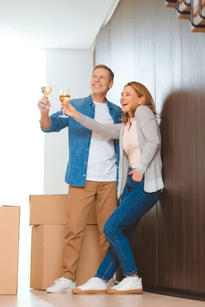 Happy couple holding glasses of white wine while standing by wall near cardboard boxes — Stock Photo