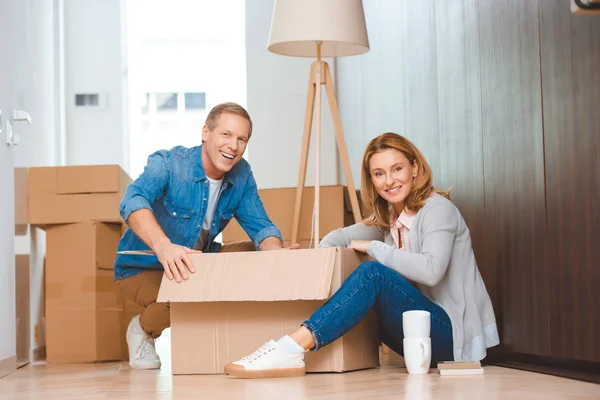 Glückliches Paar sitzt auf dem Boden und packt Karton aus — Stockfoto
