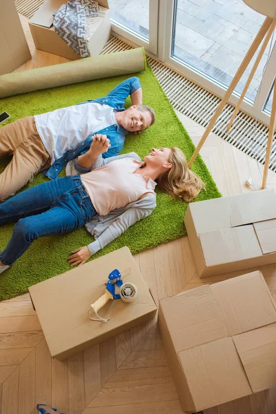 Happy couple lying on green carpet surrounded with cardboard boxes — Stock Photo