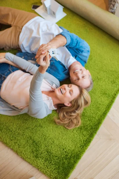 Messa a fuoco selettiva della donna che tiene chiavi con gingillo modello di casa mentre giace sul tappeto verde con marito sorridente — Foto stock