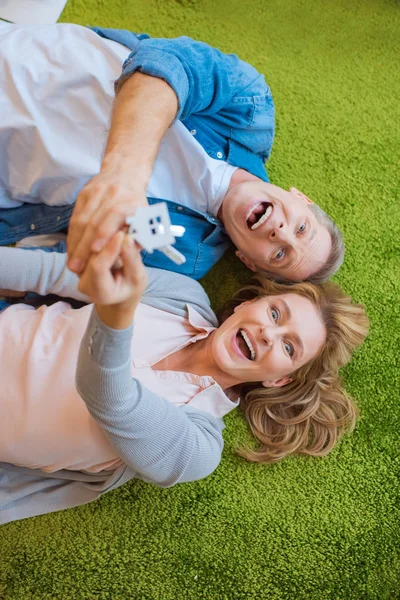 Enfoque selectivo de pareja excitada sosteniendo llaves con baratija modelo de casa mientras está acostado en la alfombra verde - foto de stock