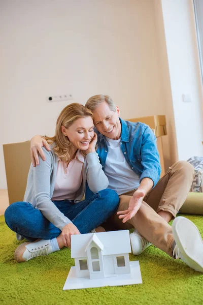 Feliz pareja abrazándose mientras está sentado en la alfombra verde cerca de modelo de casa - foto de stock