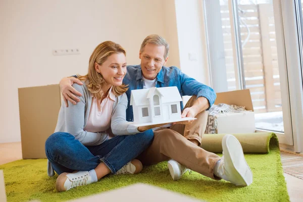 Glückliches Paar hält Hausmodell während es auf dem Boden auf grünem Teppich sitzt — Stockfoto