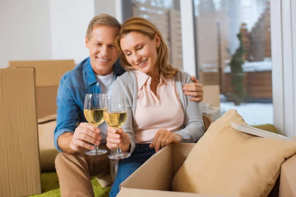 Messa a fuoco selettiva di bicchieri di vino bianco di coppia felice mentre si siede vicino a scatole di cartone a casa nuova — Foto stock