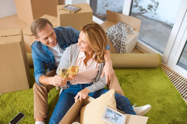 Smiling couple clinking glasses of white wine while sitting by cardboard boxes at new home — Stock Photo