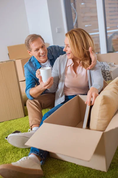 Cheerful couple unpacking cardboard box while sitting on green carpet — Stock Photo